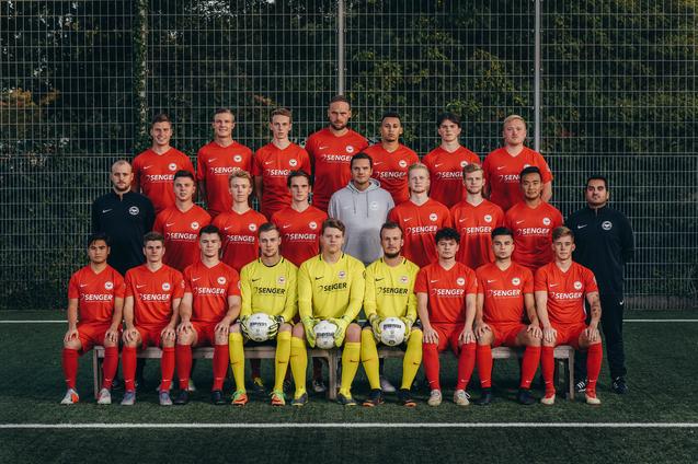 Mannschaftsfoto eines Fu&szlig;ballteams in roten Trikots, posierend auf einem Kunstrasenfeld mit zwei B&auml;llen.