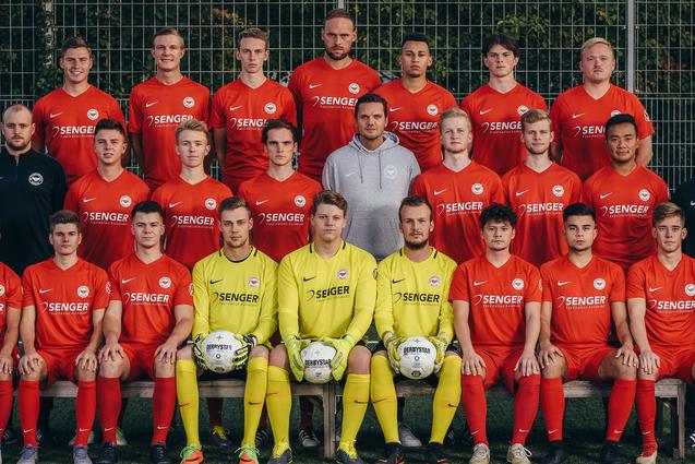 Mannschaftsfoto eines Fu&szlig;ballteams in roten Trikots, posierend auf einem Kunstrasenfeld mit zwei B&auml;llen.