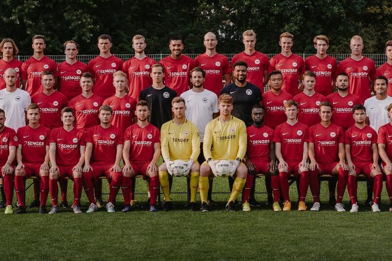 Mannschaftsfoto einer Fu&szlig;ballmannschaft in roten Trikots auf einem Rasenplatz, mit Trainern und Torwart in der Mitte.
