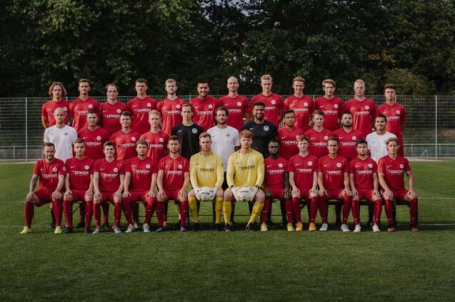Mannschaftsfoto einer Fu&szlig;ballmannschaft in roten Trikots auf einem Rasenplatz, mit Trainern und Torwart in der Mitte.