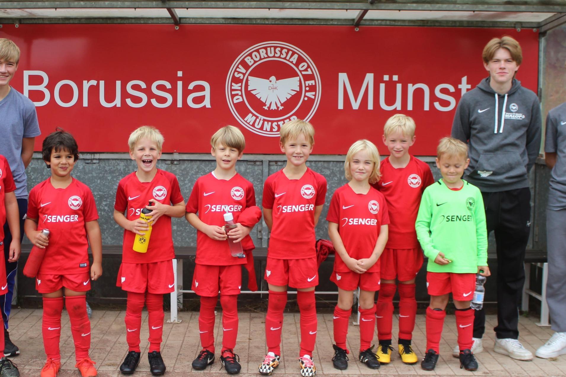 Kinderfu&szlig;ballmannschaft in roten Trikots steht l&auml;chelnd vor einem Vereinsbanner von Borussia M&uuml;nster.