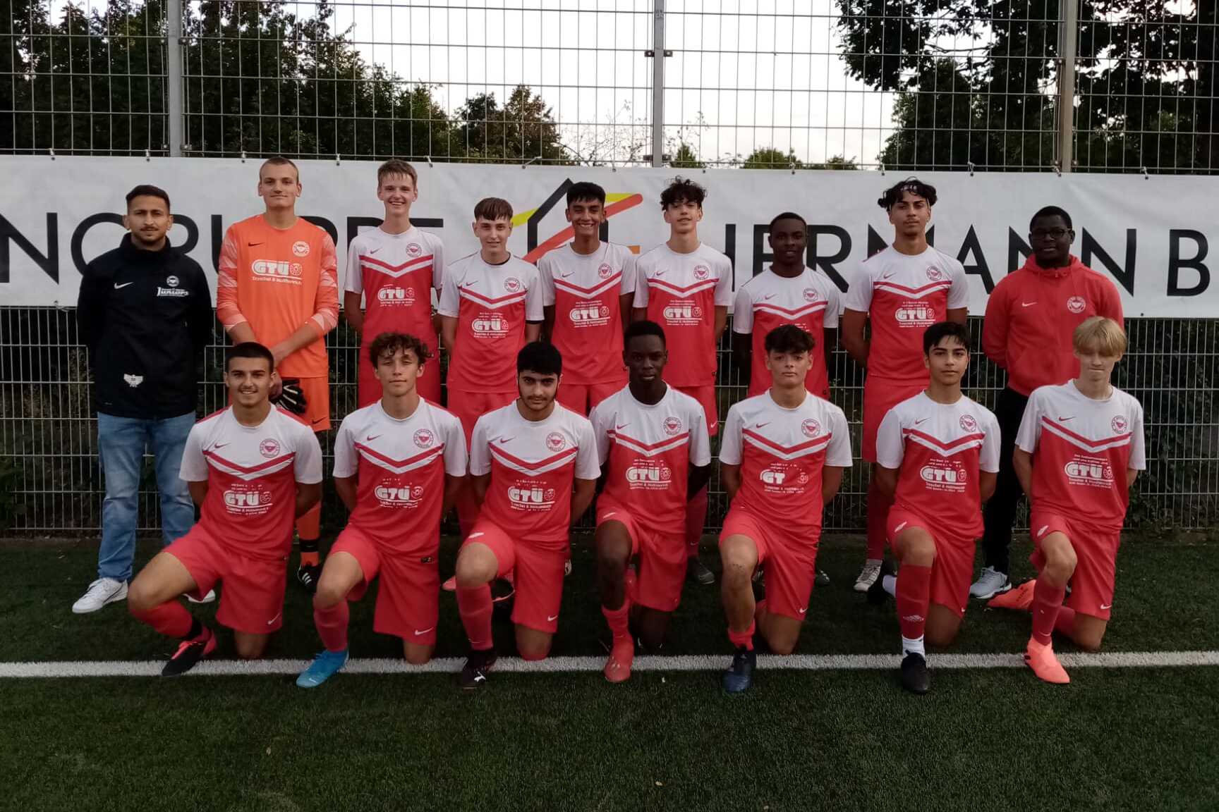 Gruppenfoto einer Jugendfu&szlig;ballmannschaft in roten Trikots auf einem Sportplatz, mit Trainer und Torwart.