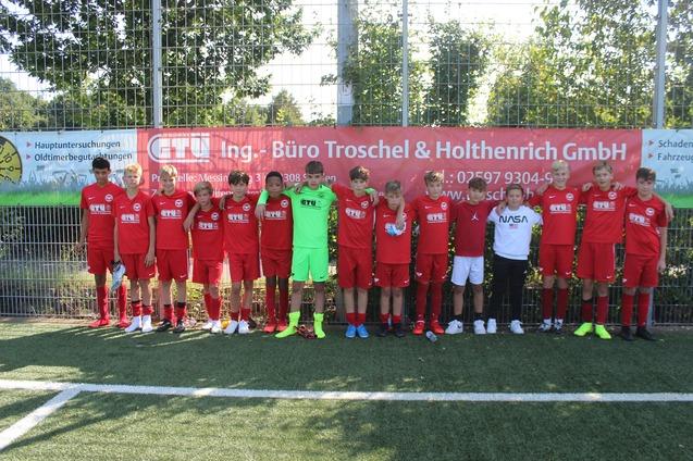 Gruppe von Fu&szlig;ballspielern in roten Trikots posiert auf einem Sportplatz mit Werbebanner im Hintergrund.