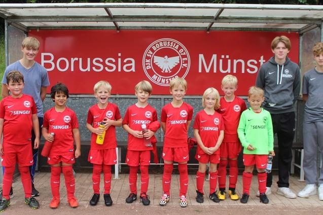 Kinderfu&szlig;ballmannschaft in roten Trikots steht l&auml;chelnd vor einem Vereinsbanner von Borussia M&uuml;nster.
