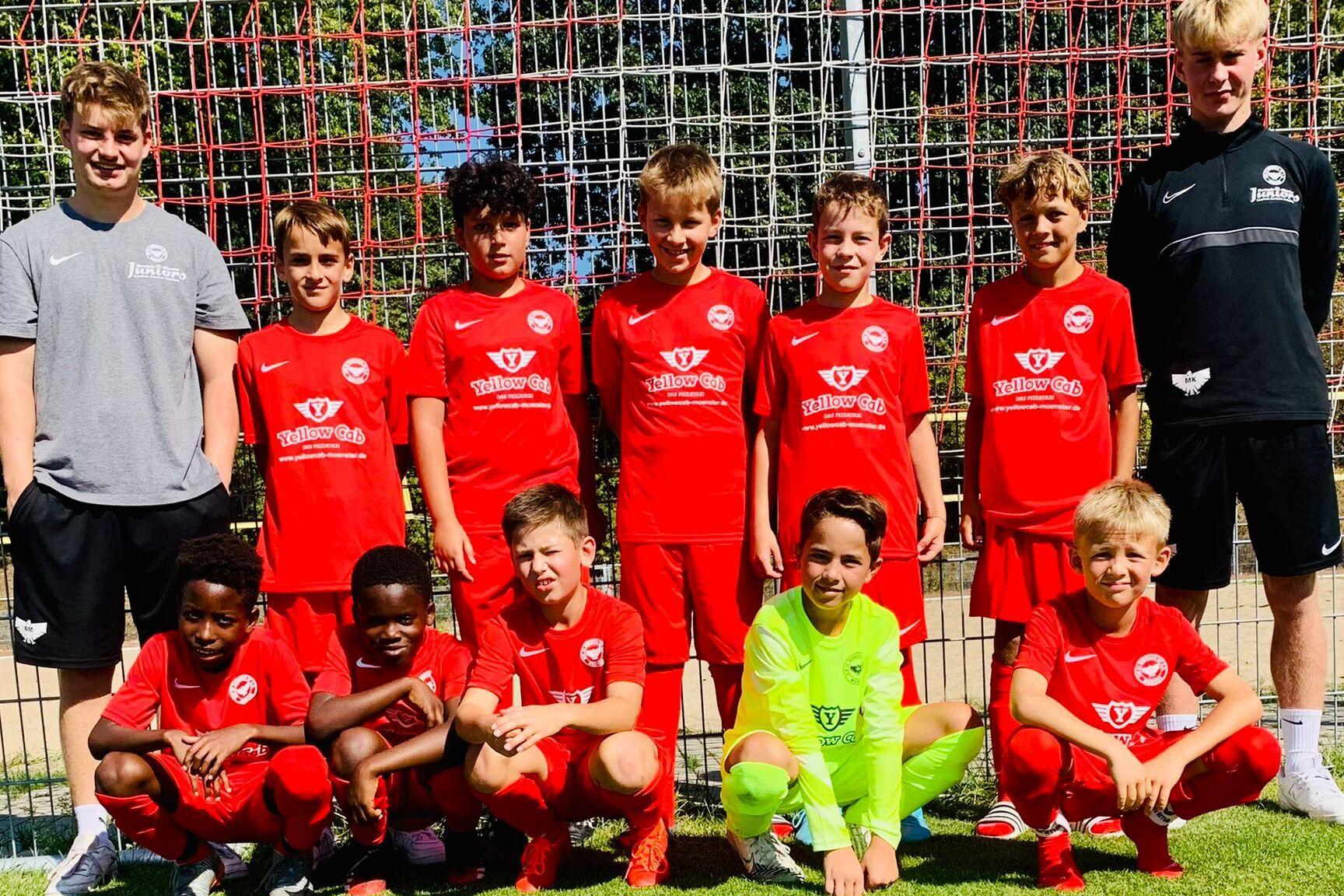 Gruppe von zehn jungen Fu&szlig;ballspielern in roten Trikots, posierend vor einem Fu&szlig;balltor auf einem gr&uuml;nen Platz.