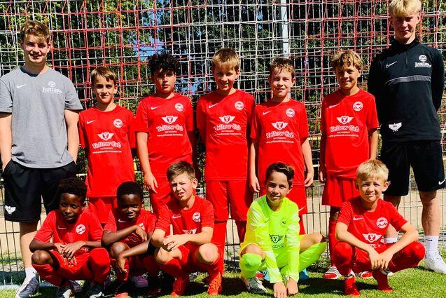Gruppe von zehn jungen Fu&szlig;ballspielern in roten Trikots, posierend vor einem Fu&szlig;balltor auf einem gr&uuml;nen Platz.