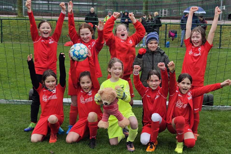 Gruppe von zehn M&auml;dchen in roten Fu&szlig;balltrikots posiert jubelnd vor einem Fu&szlig;balltor auf einem Platz.