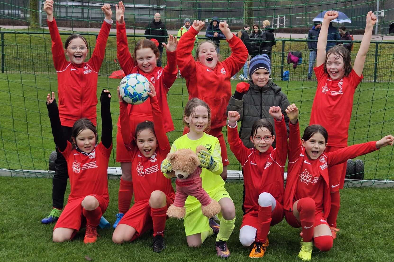 Gruppe von zehn Mädchen in roten Fußballtrikots posiert jubelnd vor einem Fußballtor auf einem Platz.