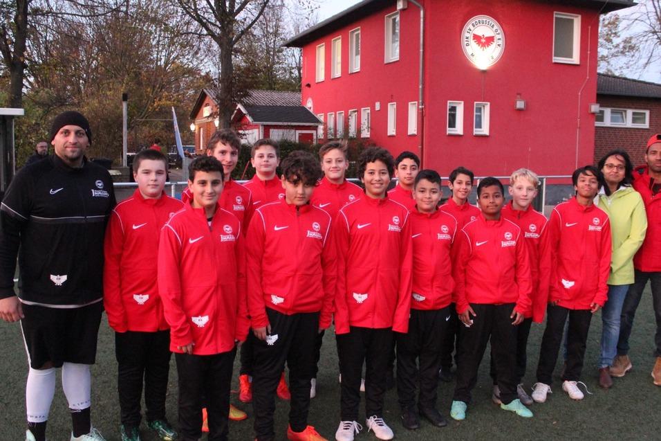 Gruppe von Fu&szlig;ballspielern in roten Trainingsanz&uuml;gen steht vor einem Sportgeb&auml;ude auf einem Kunstrasenplatz.