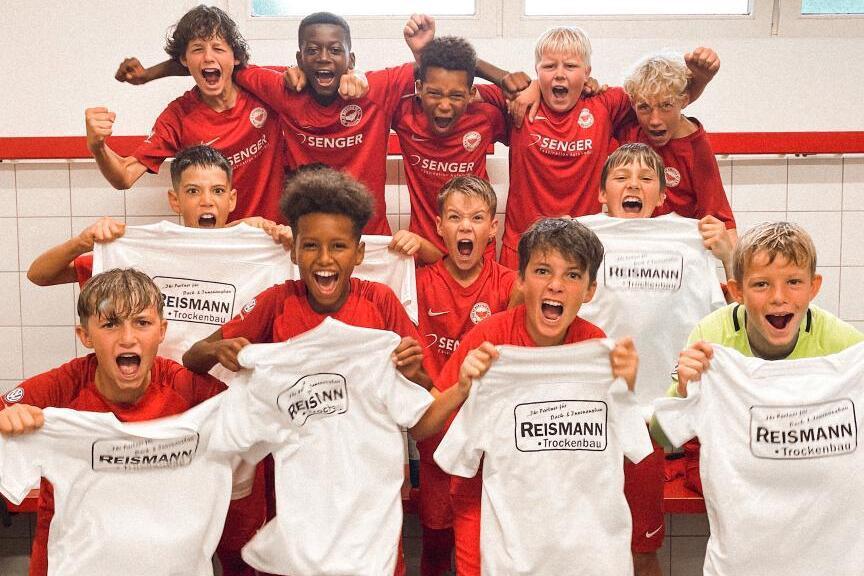 Gruppe jubelnder Kinder in roten Trikots h&auml;lt T-Shirts hoch, im Umkleideraum eines Sportvereins.