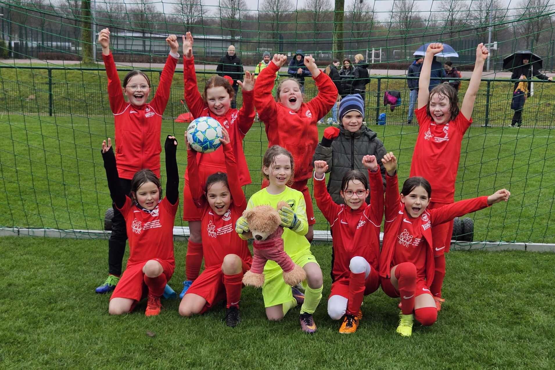 Gruppe von zehn M&auml;dchen in roten Fu&szlig;balltrikots posiert jubelnd vor einem Fu&szlig;balltor auf einem Platz.