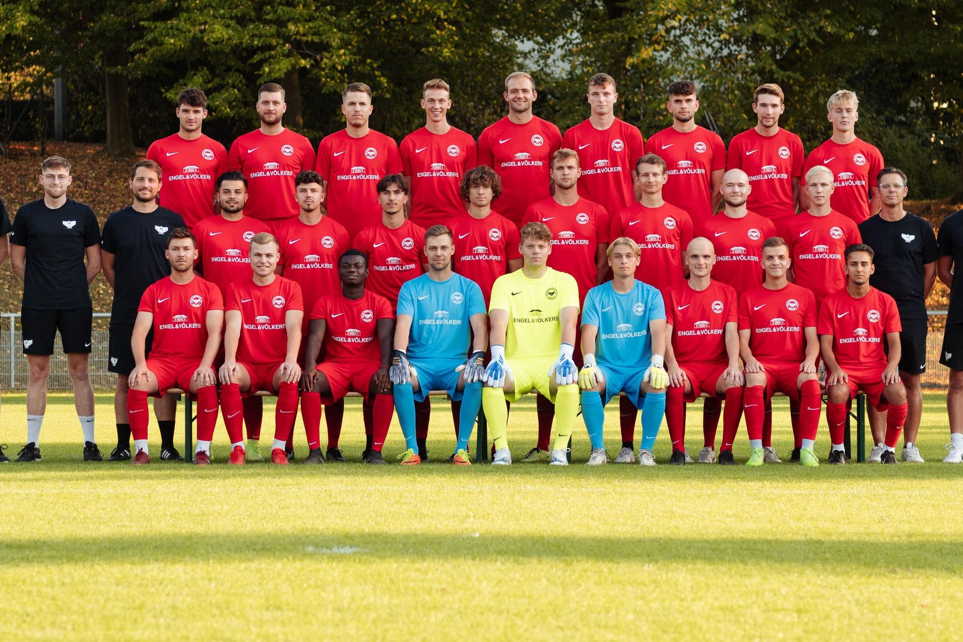 Mannschaftsfoto einer Fu&szlig;ballmannschaft in roten Trikots, gruppiert auf einem Sportplatz mit B&auml;umen im Hintergrund.