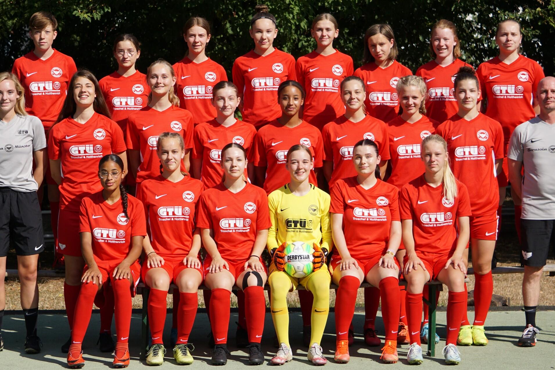 Gruppenfoto einer Fu&szlig;ballmannschaft in roten Trikots, inklusive Trainer, auf einem Sportplatz im Freien.