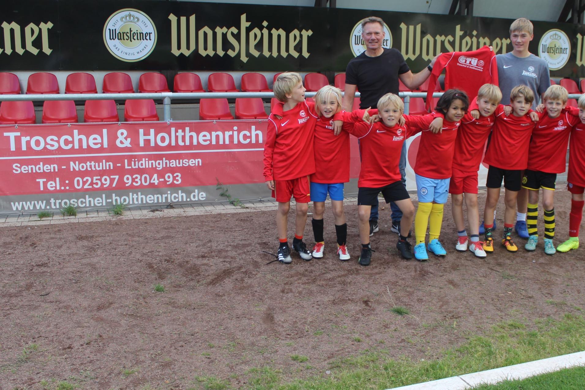Gruppe von zehn Jungen in roten Trikots posiert auf einem Fu&szlig;ballplatz, Hintergrund zeigt Zuschauertrib&uuml;ne und Banner.