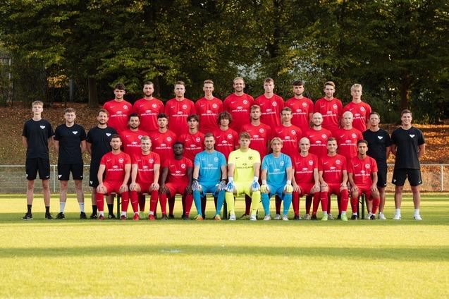 Mannschaftsfoto einer Fu&szlig;ballmannschaft in roten Trikots, gruppiert auf einem Sportplatz mit B&auml;umen im Hintergrund.
