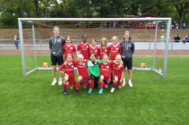Mannschaftsbild von zehn M&auml;dchen in roten Trikots und zwei Trainer:innen vor einem Fu&szlig;balltor auf einem Rasenplatz.