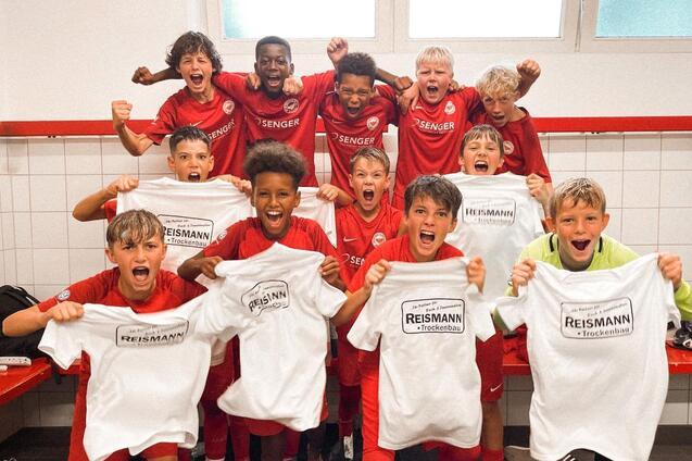 Gruppe jubelnder Kinder in roten Trikots h&auml;lt T-Shirts hoch, im Umkleideraum eines Sportvereins.