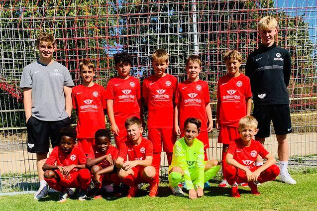 Gruppe von zehn jungen Fu&szlig;ballspielern in roten Trikots, posierend vor einem Fu&szlig;balltor auf einem gr&uuml;nen Platz.