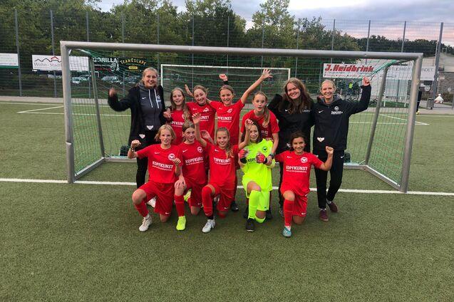 Gruppenfoto eines Fu&szlig;ballteams aus M&auml;dchen in roten Trikots, posierend vor einem Tor auf einem Rasensportplatz.