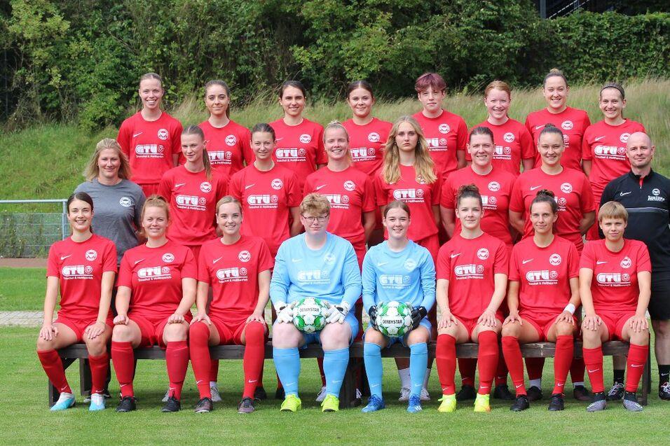 Gruppe von 24 Frauen in roten Fu&szlig;balltrikots, posieren auf einem Rasenplatz, umgeben von Gr&uuml;nanlagen.