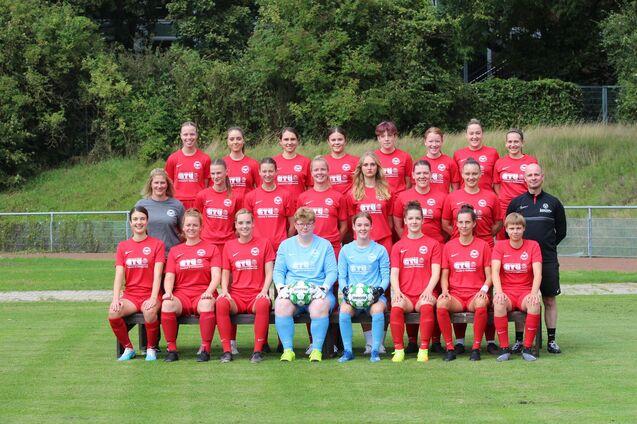 Gruppe von 24 Frauen in roten Fu&szlig;balltrikots, posieren auf einem Rasenplatz, umgeben von Gr&uuml;nanlagen.