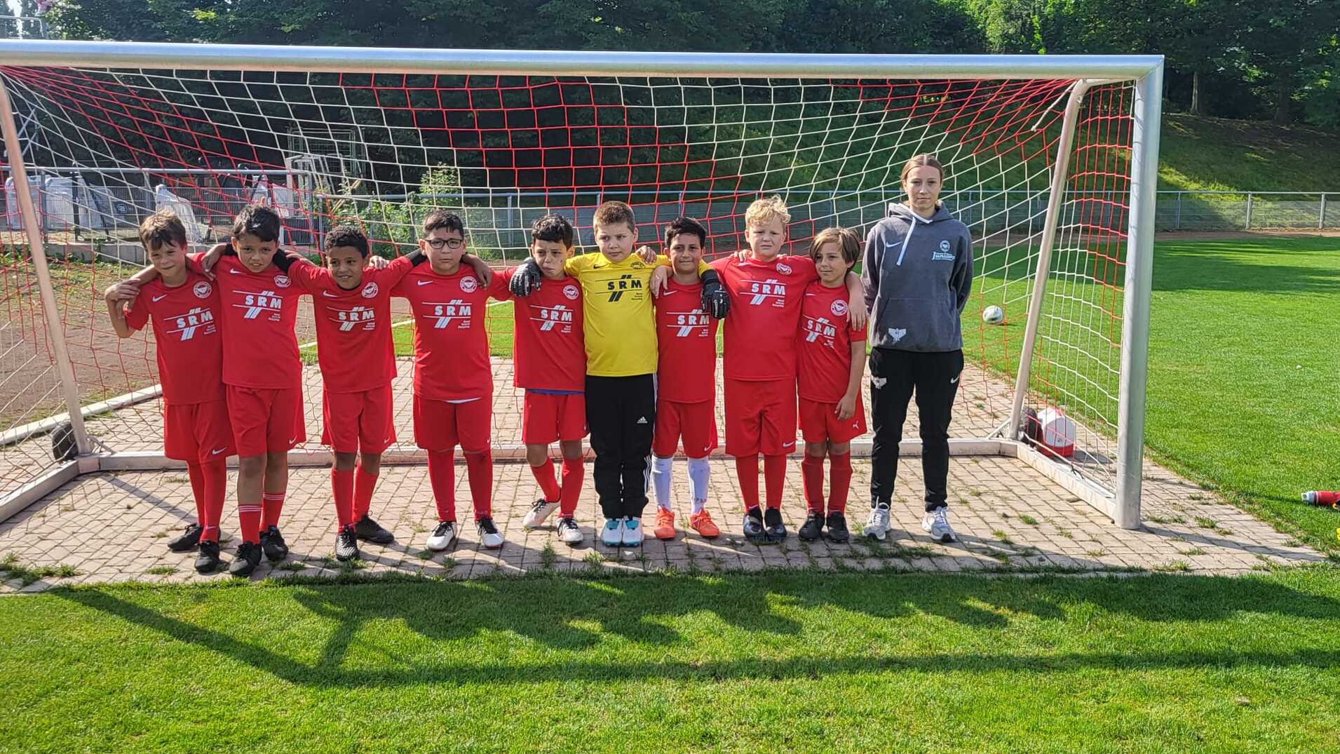 Gruppe von zehn Kindern in roten Fu&szlig;balltrikots, die vor einem Tor auf dem Spielfeld stehen, begleitet von einem Trainer.