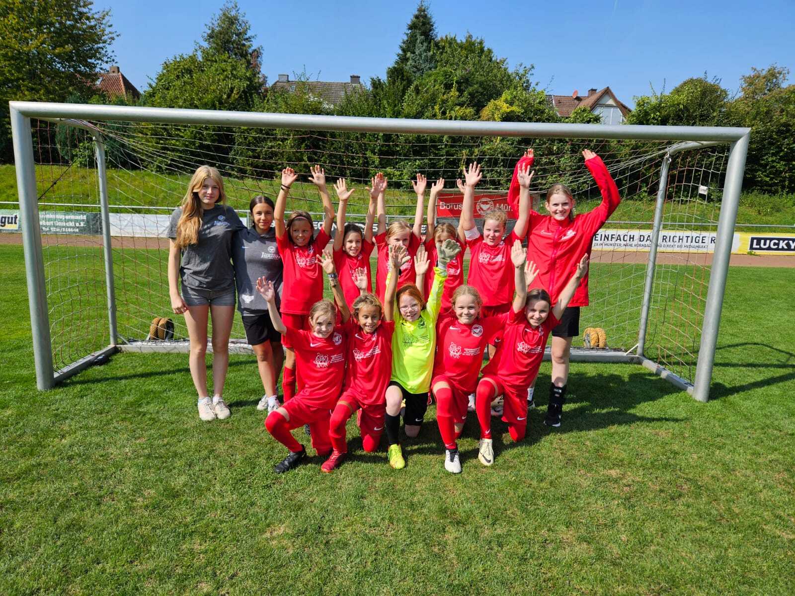 Mädchen-Fußballmannschaft in roten Trikots posiert mit erhobenen Händen vor einem Tor auf dem Rasenplatz.