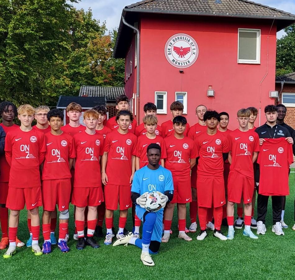 Gruppenfoto einer Jugendfu&szlig;ballmannschaft in roten Trikots vor einem roten Vereinsgeb&auml;ude in M&uuml;nster.