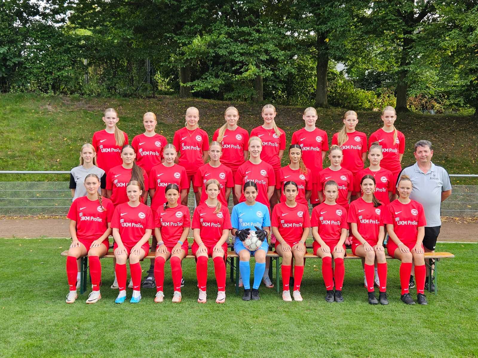 Mannschaftsfoto einer Fu&szlig;ballmannschaft mit 20 Spielerinnen in roten Trikots und einem Trainer auf einer Wiese.