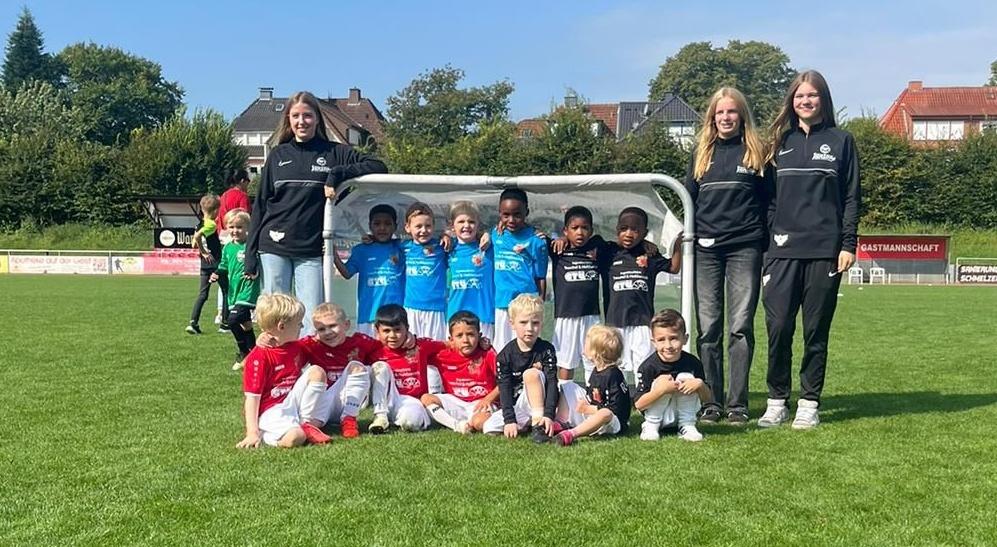 Gruppenfoto von Kindern in Fu&szlig;balltrikots und zwei Trainerinnen vor einem Tor auf einem gr&uuml;nen Spielfeld.