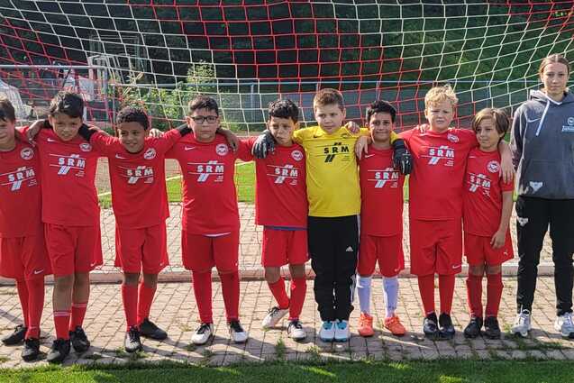 Gruppe von zehn Kindern in roten Fu&szlig;balltrikots, die vor einem Tor auf dem Spielfeld stehen, begleitet von einem Trainer.