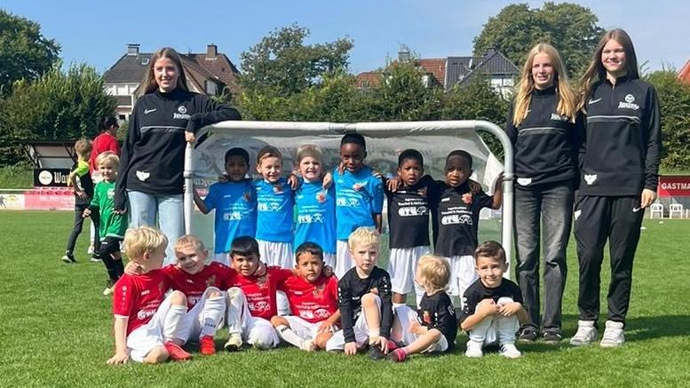 Gruppenfoto von Kindern in Fu&szlig;balltrikots und zwei Trainerinnen vor einem Tor auf einem gr&uuml;nen Spielfeld.