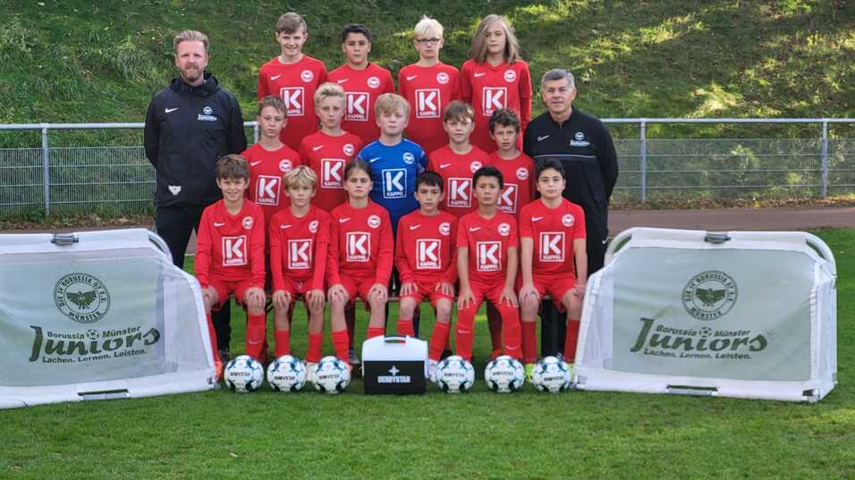 Mannschaftsbild einer Jugendfu&szlig;ballmannschaft in roten Trikots mit Trainern, B&auml;llen und Werbebannern auf dem Rasen.