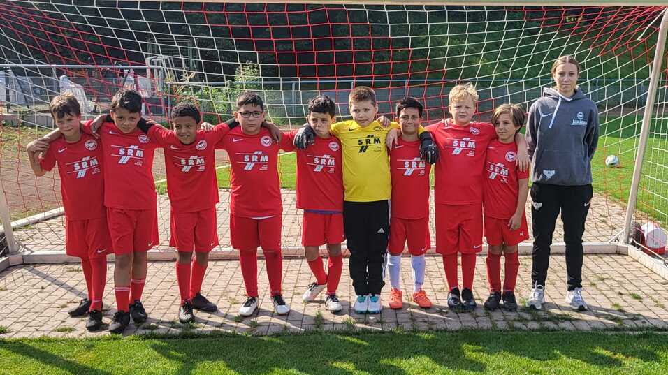 Gruppe von zehn Kindern in roten Fu&szlig;balltrikots, die vor einem Tor auf dem Spielfeld stehen, begleitet von einem Trainer.