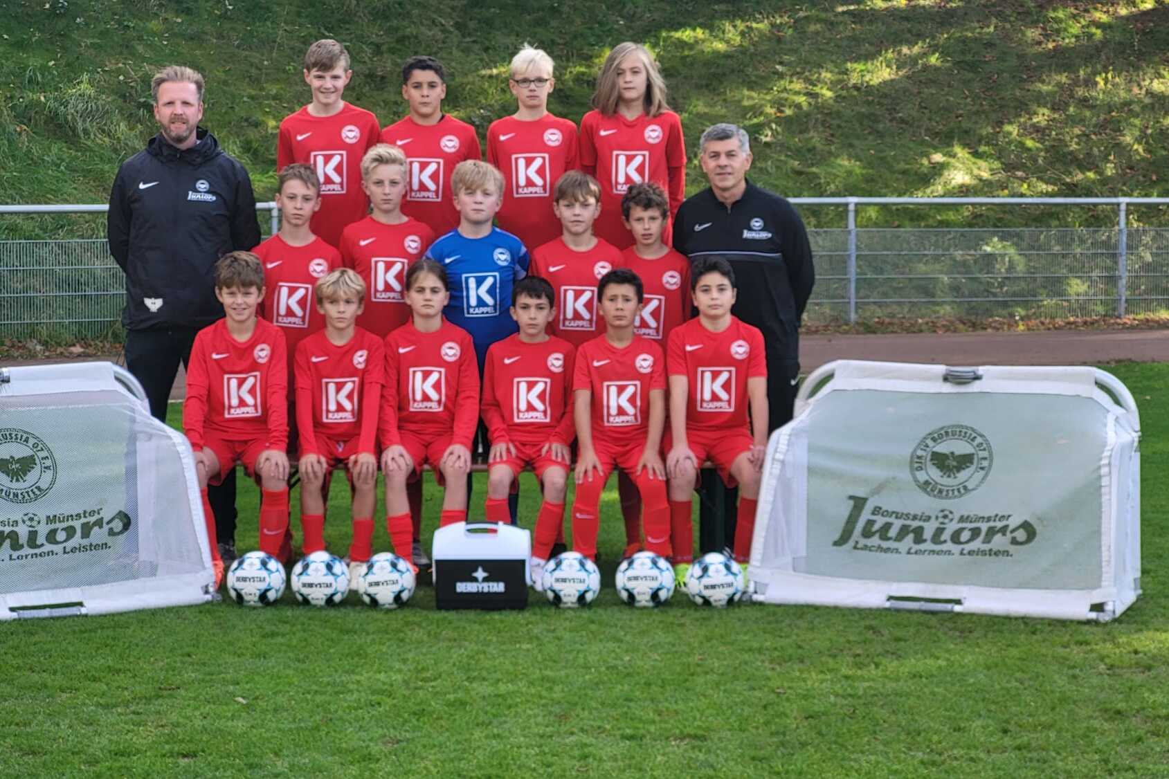 Mannschaftsbild einer Jugendfu&szlig;ballmannschaft in roten Trikots mit Trainern, B&auml;llen und Werbebannern auf dem Rasen.