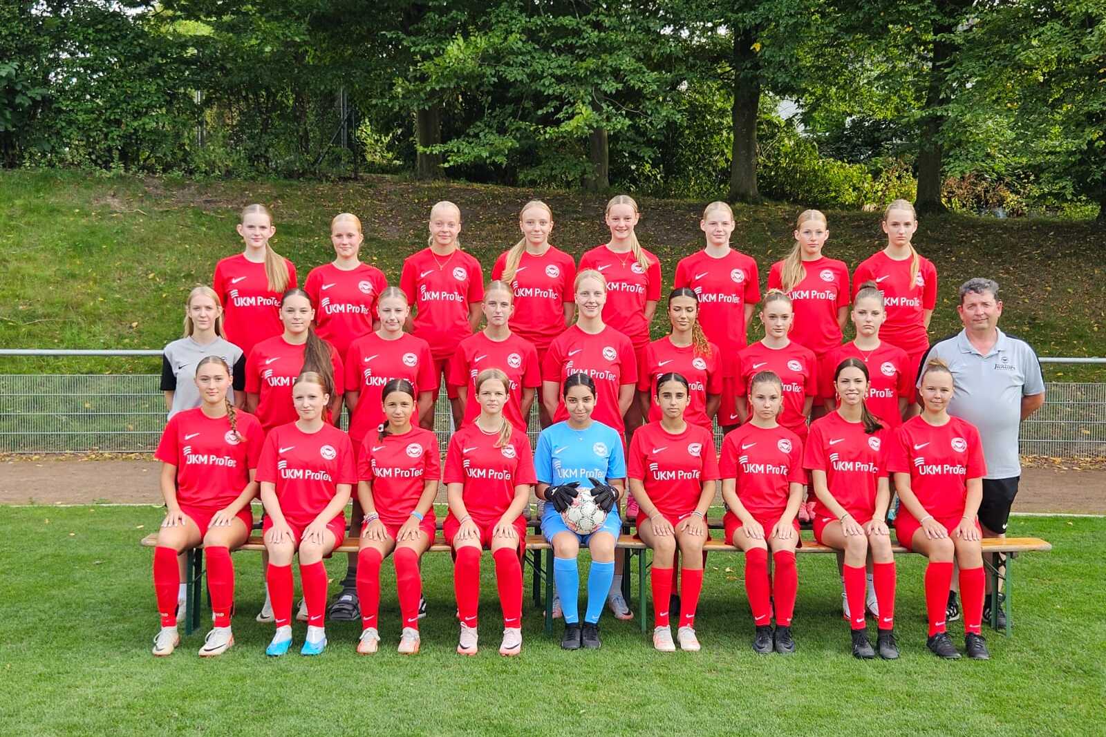 Mannschaftsfoto einer Fu&szlig;ballmannschaft mit 20 Spielerinnen in roten Trikots und einem Trainer auf einer Wiese.