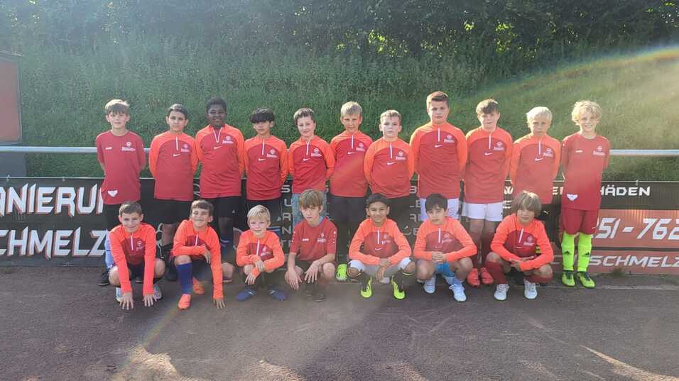 Gruppe von Jugendlichen in roten Trikots, posierend auf einem Fu&szlig;ballplatz vor einem Werbebanner.