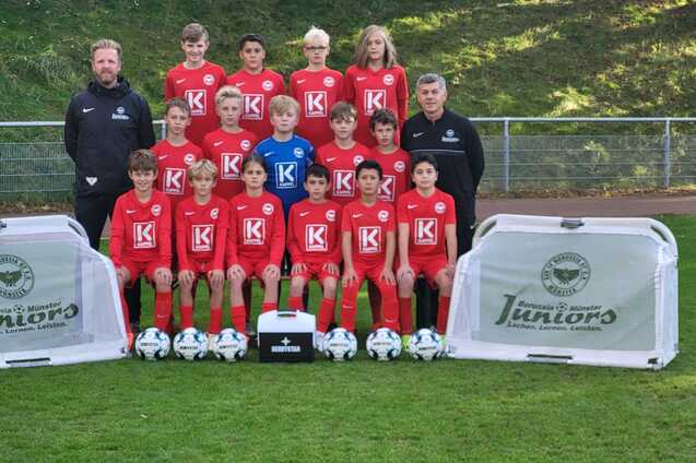 Mannschaftsbild einer Jugendfu&szlig;ballmannschaft in roten Trikots mit Trainern, B&auml;llen und Werbebannern auf dem Rasen.