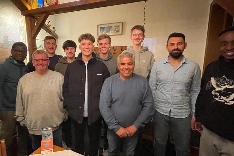 Gruppe von zehn M&auml;nnern in einer Gastst&auml;tte, l&auml;chelnd vor einer Wand mit Dekorationen und Tischdecken.