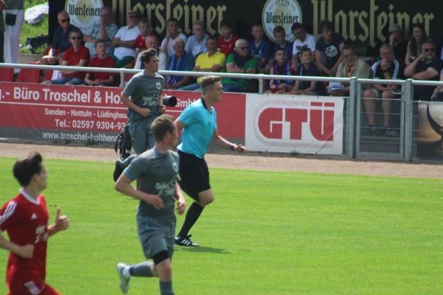 Zwei Fu&szlig;ballspieler in grauen und roten Trikots laufen auf einem Spielfeld, w&auml;hrend ein Schiedsrichter das Spiel leitet.