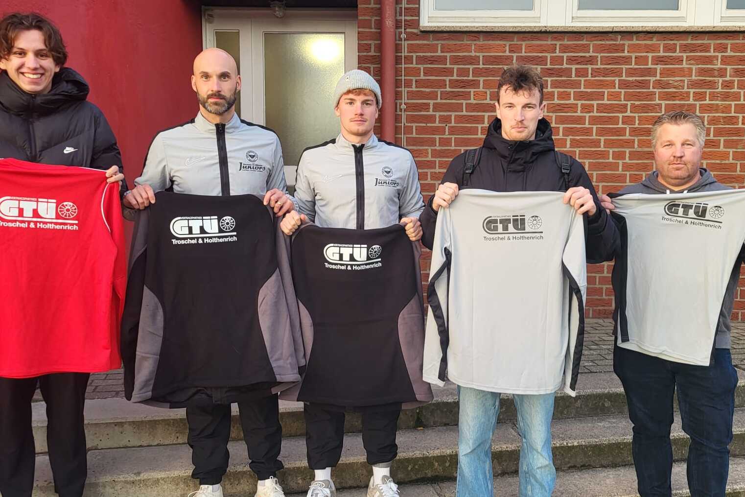 Fünf Personen stehen auf einer Treppe und halten verschiedene T-Shirts mit dem Logo „GTU“ in schwarz, rot und grau.