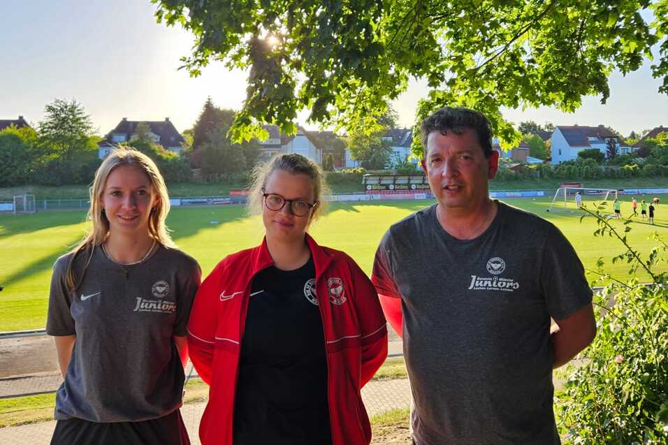 Drei Personen stehen vor einem Fu&szlig;ballfeld, umgeben von B&auml;umen bei Sonnenlicht, mit Sportkleidung und freundlichen Gesichtern.