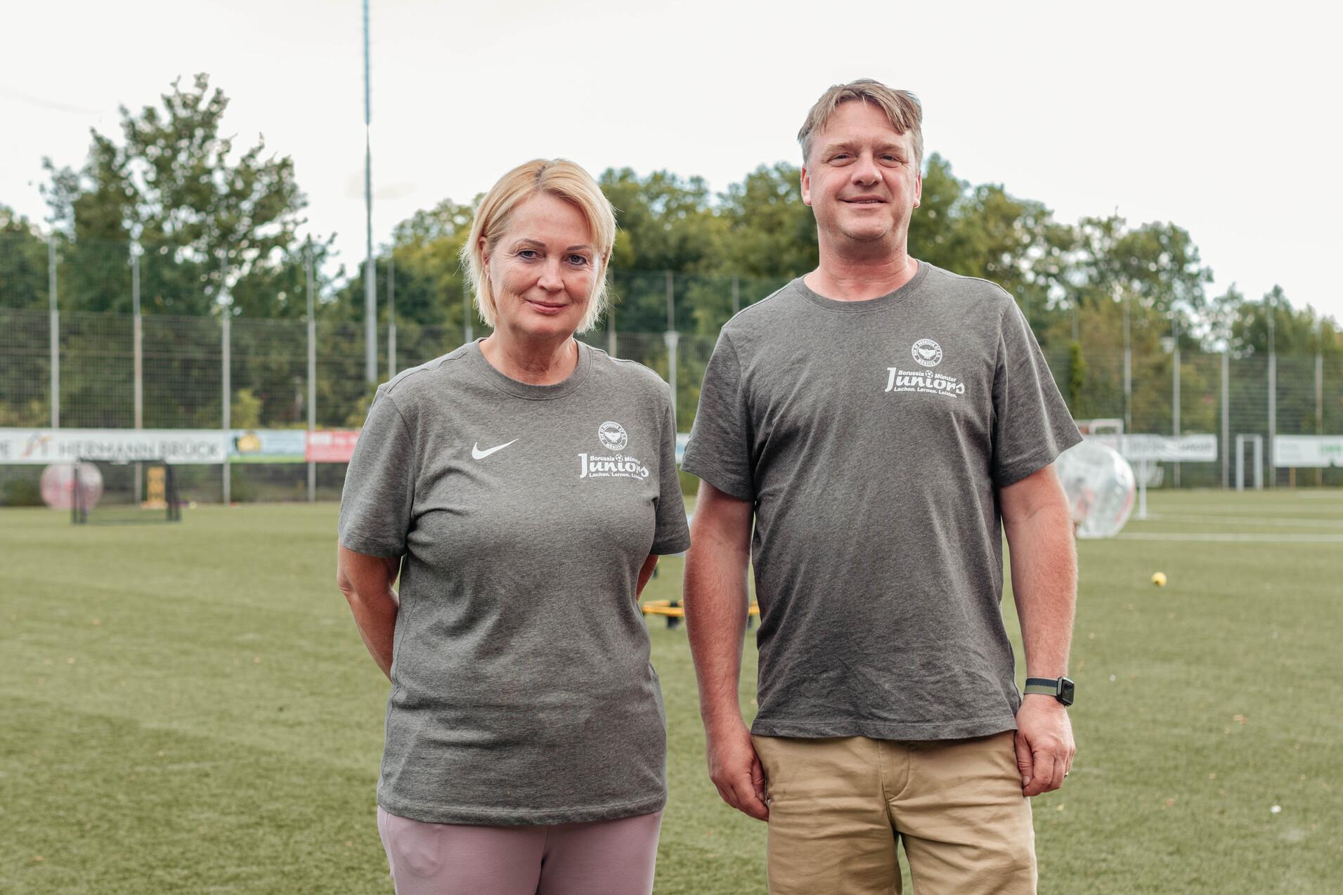 Zwei Personen in grauen T-Shirts stehen auf einem Fu&szlig;ballfeld mit gr&uuml;ner Rasenfl&auml;che im Hintergrund.
