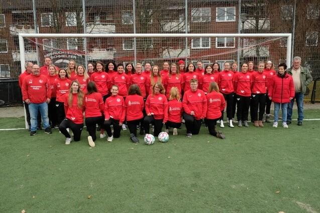 Gruppenfoto von Frauenfu&szlig;ballspielern in roten Trainingsjacken, stehend und sitzend vor einem Fu&szlig;balltor.