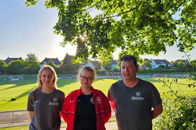 Drei Personen stehen vor einem Fu&szlig;ballfeld, umgeben von B&auml;umen bei Sonnenlicht, mit Sportkleidung und freundlichen Gesichtern.