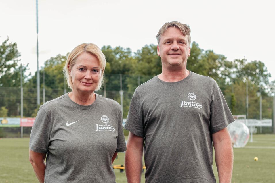 Zwei Personen in grauen T-Shirts stehen auf einem Fu&szlig;ballfeld mit gr&uuml;ner Rasenfl&auml;che im Hintergrund.