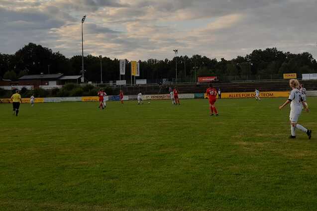 Spielszene auf einem Fu&szlig;ballplatz mit Spielern in wei&szlig;en und roten Trikots unter wolkigem Himmel.