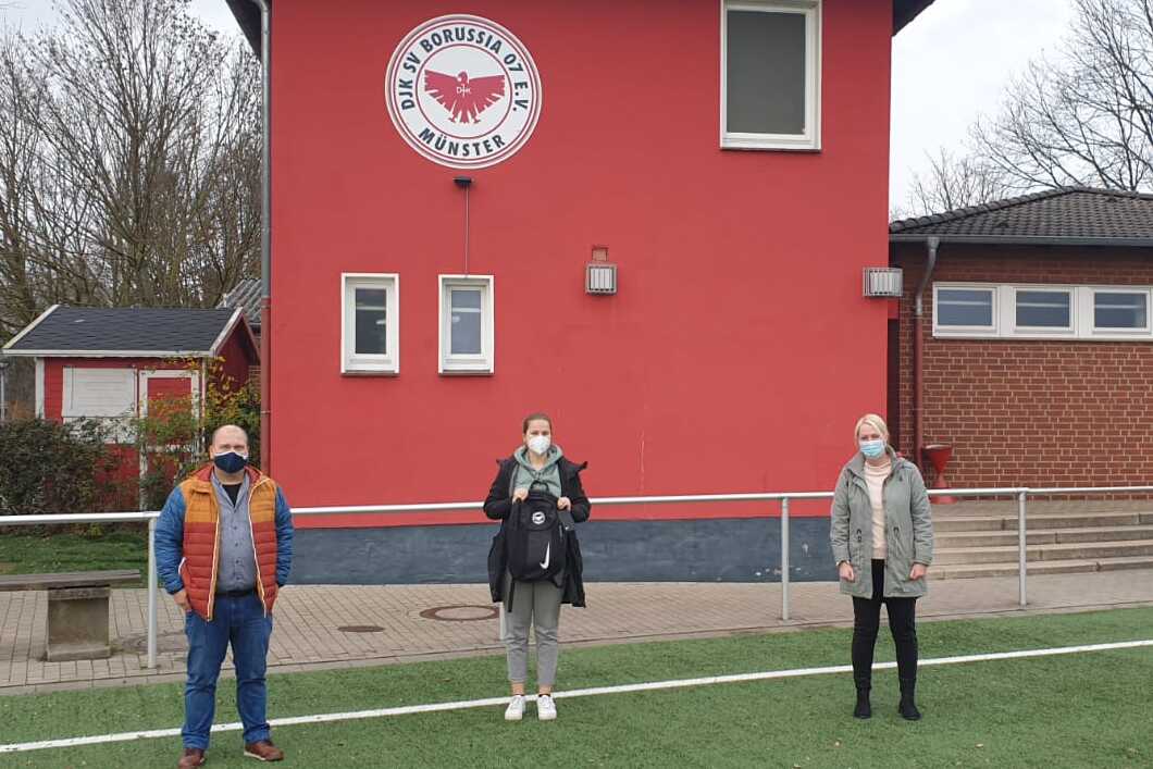 Drei Personen stehen vor einem roten Vereinsgeb&auml;ude, umgeben von einem Fu&szlig;ballfeld mit Kunstrasen.