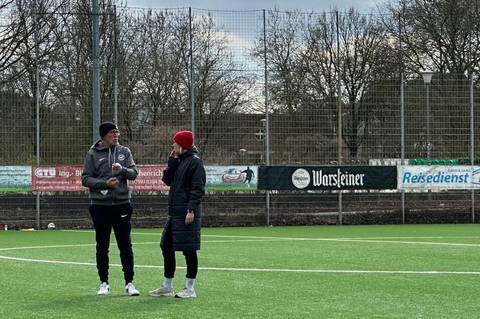 Zwei Personen stehen auf einem Fu&szlig;ballplatz aus Kunstgras und sprechen miteinander. Im Hintergrund ist ein wolkiger Himmel.