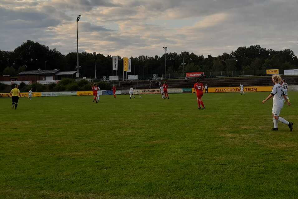 Spielszene auf einem Fu&szlig;ballplatz mit Spielern in wei&szlig;en und roten Trikots unter wolkigem Himmel.